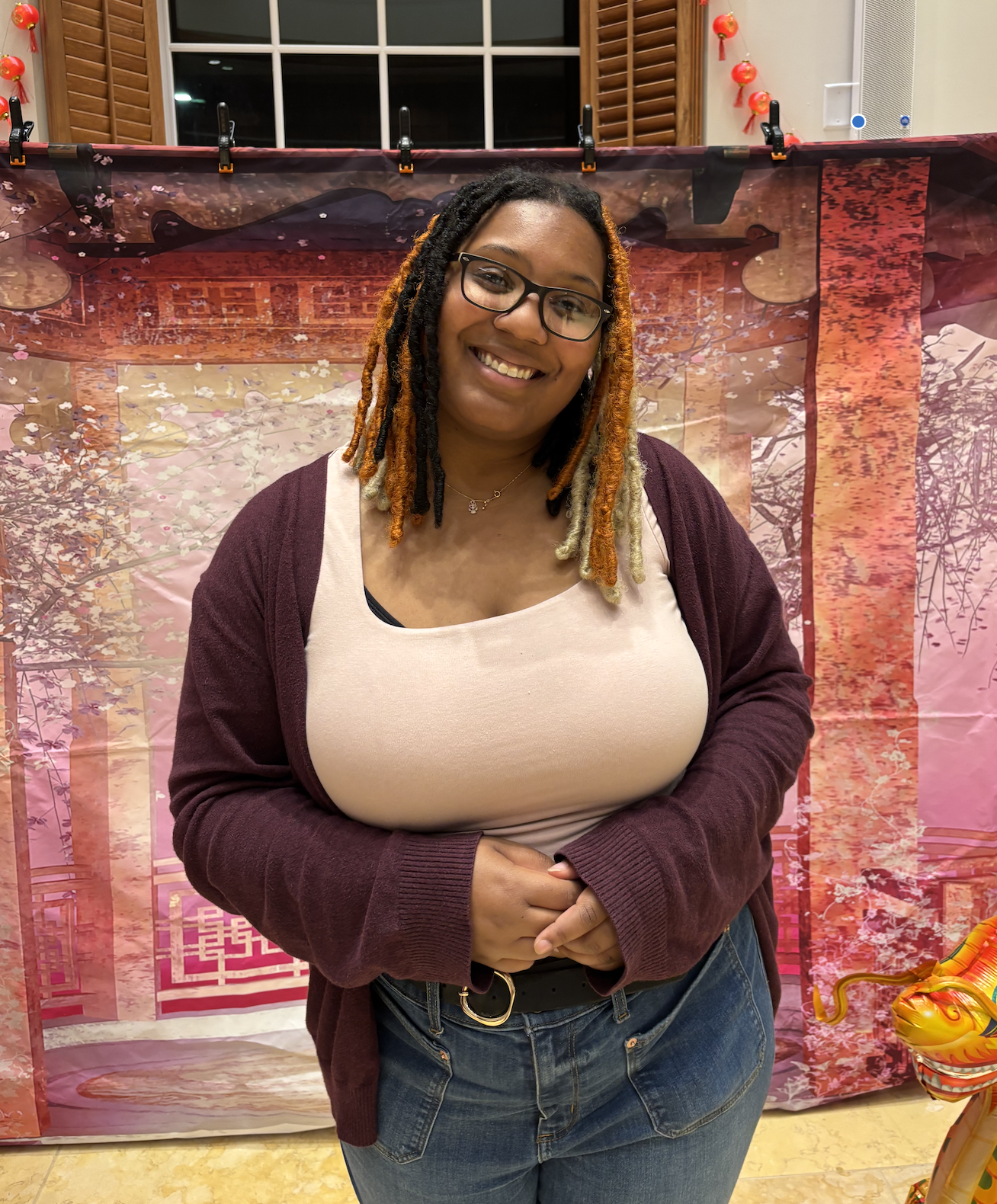 This photo features a smiling woman standing indoors in front of a decorative backdrop that appears to have an East Asian aesthetic, with imagery resembling cherry blossoms and traditional architecture. She has black and orange dreadlocks and is wearing glasses, a light beige tank top, a maroon cardigan, and high-waisted jeans with a black belt. Her hands are clasped in front of her, and she appears relaxed and happy. There are red lantern decorations hanging on both sides of the backdrop, hinting that the setting might be part of a celebration or cultural event.